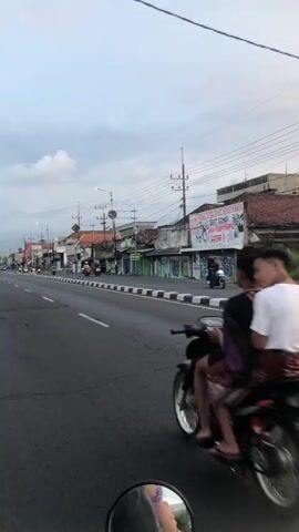 The video captures a daytime street scene with a focus on traffic. In the foreground, the perspective is from a vehicle, showing a motorcycle with two people on it riding in the same direction. The person in the back is wearing a white shirt, and the person in the front appears to be wearing a darker shirt. The motorcycle has chrome accents on its wheels. The road is paved and has a white line marking the lane. On the right side of the road, there are buildings with signs, one of which has text in red and white. There are also utility poles and wires visible. In the distance, other motorcycles and vehicles are present on the road. The sky is overcast with clouds. The overall environment appears to be a moderately busy urban or suburban street.