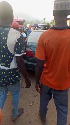 The video shows a street scene with several people walking past a parked car. The car has a large advertisement on its back window for a herbal mixture. The car is surrounded by people, some of whom are carrying objects. The background shows buildings and a slightly hilly landscape. The lighting suggests it's daytime, possibly morning or late afternoon.