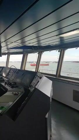 The video shows the interior of a ship's bridge. The camera pans across the bridge, revealing a large console with various instruments, monitors, and controls. There are two captain's chairs facing the console. Large windows offer a view of the sea and another ship in the distance. The ceiling is blue and made of panels. The floor is gray and reflective. The overall environment appears to be clean and well-maintained.