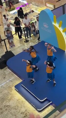 A group of four young girls, dressed in matching blue skirts and orange vests, are performing a dance routine on a blue stage. The stage is set up in what appears to be a shopping mall, with a crowd of people watching from above. A large blue backdrop with a sun graphic and text in Arabic and English is visible behind the dancers. The dancers are in various poses, some with arms outstretched and others with bent knees. The lighting is bright, and the overall atmosphere seems to be festive.