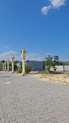 A vertical, slow-motion video pans across an outdoor scene under a bright blue sky with scattered white clouds. The foreground features a paved walkway made of grey interlocking bricks. A series of decorative, cream-colored pillars with gold accents line the left side of the walkway, leading into the distance. Behind the pillars, there are some scattered green trees and shrubs, a low grey building, and a white wall. In the middle ground, a person wearing a black shirt and a dark skirt walks away from the camera. Further in the distance, a tall transmission tower is visible on a hill. The video then shifts to focus on a large, white canopy structure providing shade over a paved area. A white car is parked in the foreground. To the right of the canopy, a dark, cube-shaped building, possibly a replica of the Kaaba, is partially visible. Another, smaller canopy structure is also present. A lamppost stands in the background. The scene suggests a public space, possibly a religious site or a rest stop.