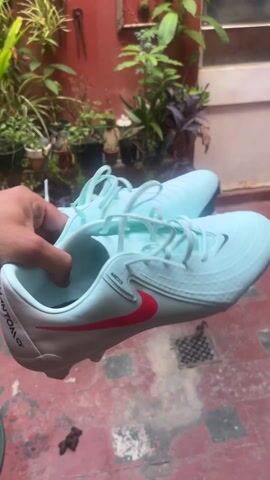 Here's a description of the video:

The video shows a person holding up a pair of light blue Nike soccer cleats. They drop the shoes, and the camera focuses on a pile of animal feces on a red-tiled patio. The patio has plants in pots, a green garden hose, and a few other items scattered around.