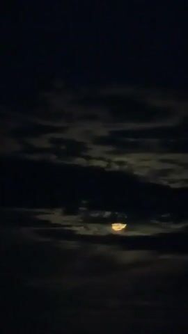 A time-lapse video shows the moon partially obscured by dark clouds. The moon appears as a crescent shape with a yellowish glow. The clouds are moving across the frame, creating a sense of depth and movement. The overall mood is serene and mysterious.