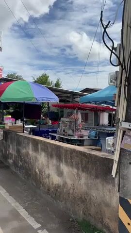 A street scene shows several food stalls lined up along a road. Colorful umbrellas provide shade for the stalls. People are standing in line at one of the stalls, suggesting it is busy. A concrete wall runs along the foreground, separating the road from the stalls. Electrical wires are visible overhead. The sky is partly cloudy.