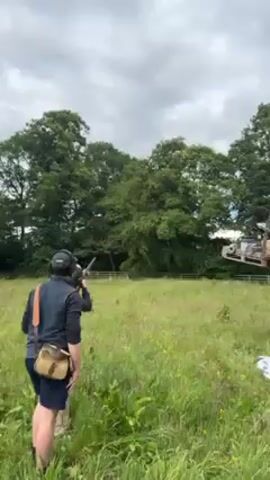 Here is a concise description of the video:

Two people are standing in a grassy field, engaged in clay pigeon shooting. One person is actively holding a shotgun, aiming and firing at a clay pigeon that has been launched into the air. The sound of the shotgun can be heard as the clay pigeon is hit and breaks apart in the sky. The scene is set in a rural environment, with a treeline and a white fence visible in the background under a cloudy sky. One person is wearing hearing protection and a vest, and the other is wearing shorts and a shoulder bag.