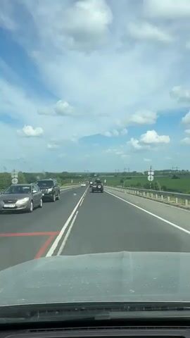 A vehicle is driving down a multi-lane highway. The weather is sunny with scattered clouds. The surrounding environment is rural, with green fields and trees. Several other vehicles are visible on the road, all moving in the same direction. The camera is positioned inside the vehicle, looking forward through the windshield.