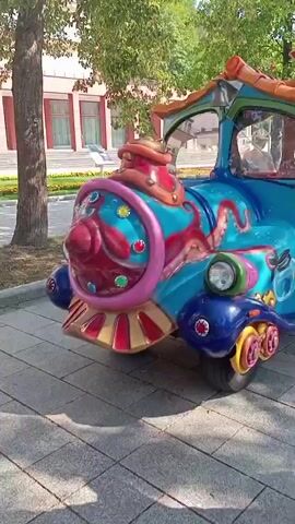 A colorful, cartoonish toy train made of blue, red, yellow, and orange plastic moves along a paved path. The train has two cars. The front car is shaped like a train engine with a light on the front. The second car is a passenger car with a roof and windows. Several children are riding in the passenger car. The train is decorated with images of sea creatures, including an octopus and a fish. The environment appears to be outdoors in a park-like setting with trees and a building in the background. The sun is shining.