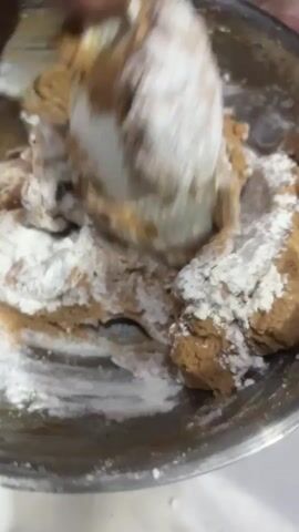 A close-up shot shows a person's hands mixing a dough in a metal bowl. The dough is a light brown color and appears to be covered in white flour. The hands are actively working the dough, using a spatula to incorporate the flour. The scene is lit, suggesting it's daytime or well-lit indoors. The setting appears to be a kitchen or food preparation area. There is no visible text. The activity is the preparation of dough, likely for baking or cooking.