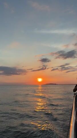 The video shows a sunset over a body of water. The sun is a bright orange orb low on the horizon, casting a golden reflection on the rippling waves. The sky is a gradient of blues and oranges, with scattered clouds tinged pink and purple. The viewpoint appears to be from the deck of a boat, with the railing visible in the lower right corner. The water is dark and choppy, creating a wake behind the boat. A distant landmass is visible on the horizon, silhouetted against the setting sun.