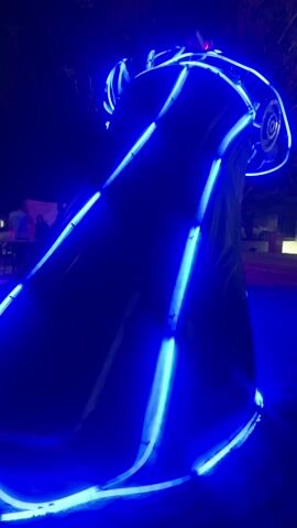 At night, an oddly-shaped structure glows with brilliant blue neon lights. The structure is dark underneath its linear neon highlights. At its top is what appears to be a speaker. In the distance, there are a few people sitting at a table, and someone walking by.