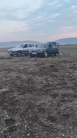 The video begins with a close-up of two vehicles parked in a muddy, open field. The vehicle on the left is a silver SUV, and the vehicle on the right is a black sedan. In the background, mountains are visible under a cloudy sky with hints of pink and orange from a sunset. As the video progresses, the camera pans slowly to the right, revealing more of the field. Scattered across the ground are various objects, including what appear to be bags, bottles, and possibly blankets, suggesting a gathering or picnic. The camera then pans back to the left, showing the two cars again. The scene appears to be outdoors during twilight, with the light fading. No people are clearly visible in the frame.