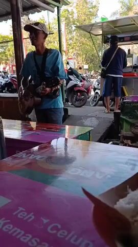 A man wearing a blue t-shirt, denim jeans, and a beige baseball cap stands holding a guitar and appears to be singing. He is positioned in front of several colorful tables with laminated surfaces. Behind him, a second person, wearing a dark jacket and blue shorts, stands with their back to the camera, seemingly interacting with a food stall. Several motorcycles are parked in the background, and trees are visible beyond. The scene appears to be an outdoor marketplace or food court.