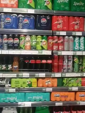 The video shows a grocery store refrigerator aisle. The refrigerators are glass-fronted and contain shelves stocked with various beverages and dairy products. On the first shelf from the top in the left refrigerator, there are blue packs of Pepsi. Below that are more Pepsi products, and then green and orange bottled drinks. The bottom shelves are stocked with more drinks in blue and green packaging. In the right refrigerator, the shelves are filled with milk jugs and other dairy products. Two men are visible in the aisle. One man in a green shirt is reaching into the refrigerator, while the other man in a blue shirt is standing and looking at the shelves. The overall environment appears to be clean and well-stocked.