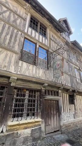 The video pans across the exterior of several old, timber-framed buildings. The architecture suggests a historic European town. The street is paved with cobblestones. A sign hangs from one building, indicating a museum or gallery. A single person, wearing a light-colored coat, walks away from the camera down the narrow street. The sky is blue with scattered clouds. The overall impression is one of a quaint and ancient urban setting.