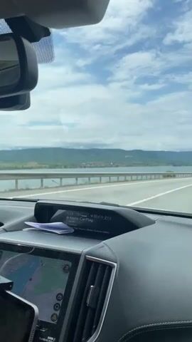 The video captures a first-person view from inside a moving car, looking out through the windshield at a scenic landscape. The car's interior features a dashboard with an infotainment screen displaying "Apple CarPlay", "19:33", "19:0", and "21°C". A smartphone mounted on the dashboard shows a navigation map. A rearview mirror is visible at the top.

Outside the car, a multi-lane asphalt road with white markings runs alongside a metal guardrail. Beyond the guardrail, a wide body of water, likely the Danube River given the location context of Drobeta-Turnu Severin, Romania, stretches into the distance. Across the water, rolling green hills and mountains are visible, dotted with houses forming a small town or village. The sky is partly cloudy with patches of blue.

The activity involves driving along this road, enjoying the scenic views. The time of day appears to be daytime, with bright but overcast weather. Music is playing in the background throughout the video, contributing to the ambiance of a road trip.