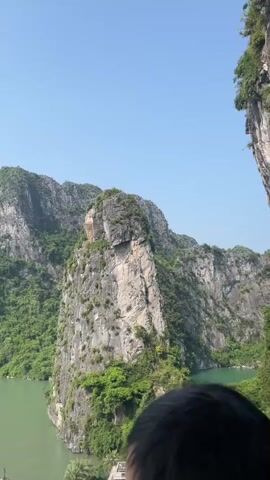 The video opens with a close-up shot of a large, steep, rocky island covered in lush green vegetation. The island is surrounded by calm, green water. A small portion of a person's head is visible in the lower right corner of the frame. The camera then pans slowly to the right, revealing more of the island and the surrounding seascape. Several boats are visible in the water, including a large tour boat in the foreground and smaller boats in the distance. The sky is clear and blue, with a hazy horizon. The video continues to pan to the right, showing a bay filled with numerous tour boats navigating between more of the iconic limestone karsts that characterize the region. The scene is serene and showcases the natural beauty of the landscape.