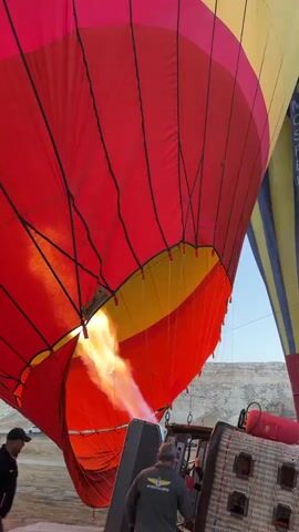 A close-up shot shows the bottom of a red and yellow hot air balloon being inflated by a propane burner. The burner, positioned directly beneath the balloon's opening, emits a large orange flame that illuminates the fabric of the balloon. Two individuals are visible in the foreground. One, wearing a red jacket and a cap, stands to the left, observing the process. Another person, wearing a grey jacket and a cap, is positioned near the burner. The background reveals a second, partially visible hot air balloon with yellow and blue sections, and a dry, arid landscape under a bright sky. The scene appears to be an outdoor preparation area for a hot air balloon launch.