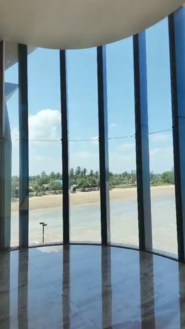 The video shows a panoramic view of a beach and the ocean through a curved window with vertical frames. The sky is blue with some white clouds. A sandy beach with some greenery and a few scattered structures is visible. The water is a light brown color with small waves. The interior of the room, which has a polished floor, is reflected in the window.