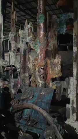 The video displays a close-up view of an abstract sculpture constructed from various materials, including what appears to be aged metal. The sculpture has a rough, textured surface with patches of rust and verdigris, suggesting significant weathering or an intentional patina. Several elements are visible within the sculpture, including a watch on the left side and what looks like a relief of an animal, possibly a snake or lizard, on a metallic surface in the foreground. The background is dark and indistinct, implying an indoor setting, possibly a workshop or exhibition space. The lighting is low, highlighting the details and textures of the sculpture.