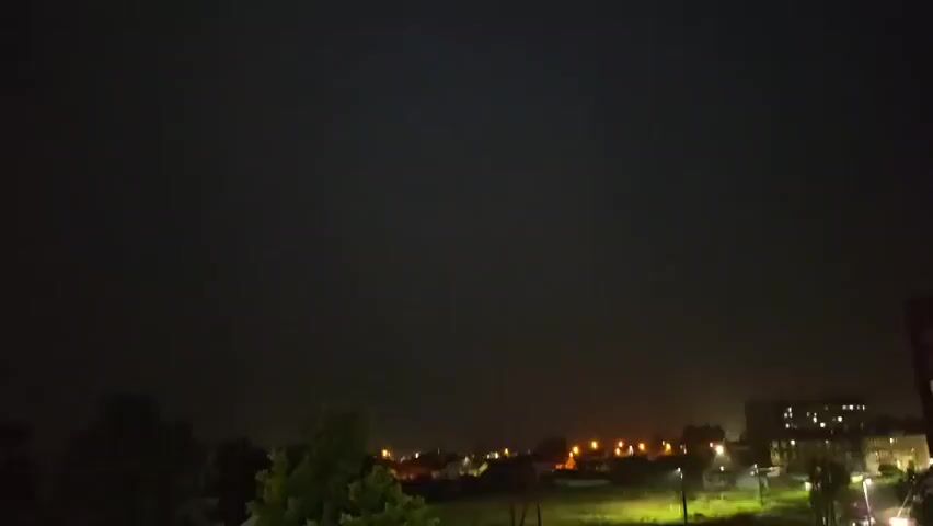 A night sky is shown, with a distant city illuminated by streetlights and building lights. Trees are visible in the foreground. There is no audio.