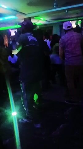 The video shows a dimly lit nightclub with a mix of colorful, mostly red and green lighting. A small group of people are gathered, some standing and some moving, obscured by the low light. One person is wearing a white cap and a plaid shirt. Another person is wearing dark clothing with white stripes down the side and the word "pama" on the back. The floor is dark and shiny, reflecting the lights. A decorated column is visible on the right side of the frame. The overall atmosphere is active and social.