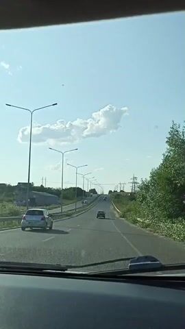 The video shows a view from inside a car driving on a highway. The sky is blue with a few scattered clouds. There are streetlights along the side of the road and power lines in the distance. Several cars are on the highway ahead. The road is paved and appears to be in good condition. Trees and greenery are visible on the sides of the road. The dashboard of the car is visible in the foreground.