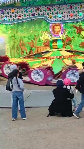 The video shows a carnival ride with several people in the foreground. In the middle ground, the ride is a circular attraction with multiple cars that spin and move. The ride is brightly lit with flashing lights of various colors, including blue, pink, and red, and features images of what appear to be movie stars or performers on its backdrop. The ride is in motion, with the cars blurring due to their speed. In the foreground, three individuals are standing and observing the ride. Two young girls and a woman are facing the ride. One girl is wearing a grey hoodie and blue jeans, and the other is wearing a black jacket and a pink helmet. Another person, possibly an adult, is kneeling behind them, embracing the girl with the pink helmet. To the left, a woman wearing a white headscarf and grey hoodie, and blue jeans, is standing and holding a phone, appearing to take a picture or video of the ride. The ground in the foreground is a sandy or gravelly surface. The atmosphere suggests a lively and festive environment typical of a fair or amusement park.
