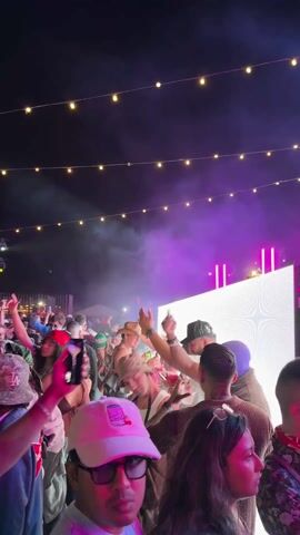 The scene shows a crowd of people at an outdoor event at night. The sky is dark and there are string lights overhead. In the background, there is a stage with a bright white screen and purple lighting. Smoke or fog is being released. Many people are facing the stage, some with their arms raised and holding up phones. The crowd seems to be enjoying a concert or party.