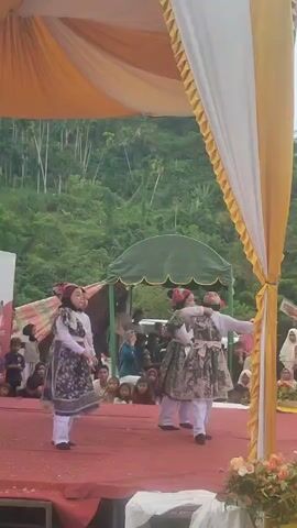 The video captures a stage performance where three young girls in traditional attire are dancing. The stage is adorned with red carpet and decorated with white and yellow drapes. In the background, a lush green hill covered with trees is visible. An audience is seated behind the stage, watching the performance. The girls are dressed in colorful outfits, including headscarves and patterned robes. They appear to be performing a coordinated dance routine, with one girl in the front raising her hands, while the other two move in unison. The lighting suggests it is daytime, possibly an outdoor event.
