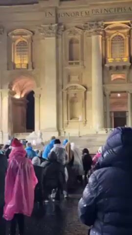 In a night-time exterior setting, a large group of people stands, mainly facing away from the camera, in what appears to be a public square. Most wear rain gear, including ponchos and hooded jackets in various colors like blue, pink, and black. They are watching a large screen that displays an ornate interior, likely religious, possibly a cathedral. Above the screen is a statue of a man, presumably a historical figure, and the building behind the statue is illuminated, featuring architectural details like pillars, arched entrances, and a row of statues atop the roofline. The words "IN HONOREM PRINCIPE" are visible on the building. The atmosphere suggests a public gathering, possibly for a religious event or viewing. The weather appears to be rainy or damp, given the rain gear worn by the attendees.