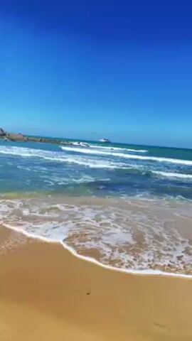The video shows a sunny beach with golden sand and turquoise water.  Waves are crashing on the shore. In the background, there is a mountain range covered in greenery. The sun is bright, and there are some people visible on the beach, though they are small and distant.  The overall view is a scenic coastal landscape.