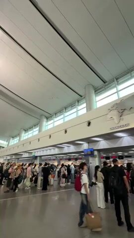 The video shows a long queue of people waiting in an airport terminal. The ceiling is high and white, with a world map depicted on the wall above the check-in counters. People are standing in lines, some with luggage, facing the counters. The lighting is bright and airy, suggesting a daytime scene. The atmosphere appears to be busy but orderly.