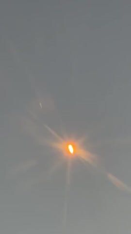 A short video clip shows a bright orange, circular object in the sky, possibly the sun, with bright rays emanating from it. The background is a hazy, pale blue sky. There are lens flares present in the video, suggesting it was filmed through a camera lens. The object appears to be stationary or moving very slowly.