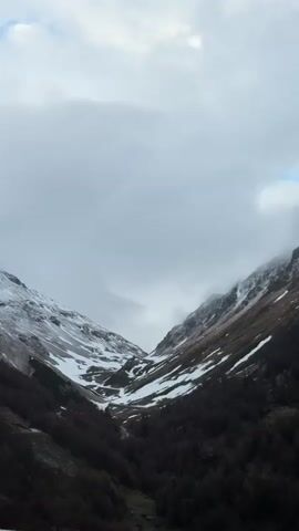 The video captures a wide, scenic view of a mountain valley in Poschiavo, Switzerland, under an overcast sky. The camera slowly pans from right to left, revealing the expanse of the landscape. Steep mountain slopes, partially covered with patches of snow, flank both sides of the deep valley. The lower sections of the mountains are densely forested with dark, possibly coniferous or bare deciduous trees, suggesting late autumn or early spring.

The sky is filled with heavy, grey clouds that hang low, partially obscuring the higher peaks and creating a diffused, soft light. Wisps of mist or lower clouds can be seen clinging to the upper slopes, adding to the dramatic atmosphere. In the foreground, the terrain is covered with dry, brownish grass, indicative of the season. No people or animals are visible, and the scene exudes a sense of serene, rugged wilderness. There is no discernible text in the video.