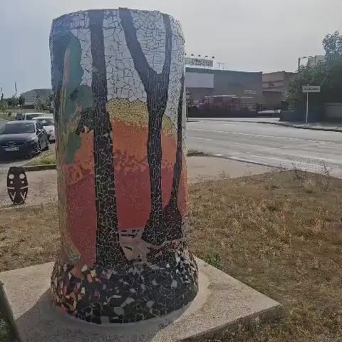 A cylindrical mosaic sculpture depicting trees and a sunset is displayed on a concrete base. The sculpture is located outdoors, with a street and buildings visible in the background. Several cars are driving on the street, and some pedestrians are walking on the sidewalk. The sun is shining, and the sky is clear.