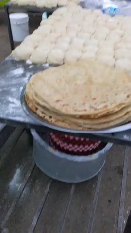 Here is a description of the video:

The video shows an outdoor food stall with a stack of already-cooked chapati bread in the foreground. Behind the stack, there's a table covered in dozens of dough balls. To the right, a man stands at a charcoal grill, cooking chapatis, periodically flipping them with a spatula. Other people are seen in the background, moving about, suggesting a busy location. A street scene is visible in the distance. Various advertisements are placed in the background, indicating the presence of nearby businesses.