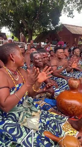 A group of women are seated and clapping their hands. They are dressed in colorful, patterned fabric and some are wearing jewelry. In the foreground, there are several gourds and a musical instrument. In the background, there are more people watching, some tents, and trees. The women appear to be participating in a cultural event or ceremony.
