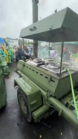 The video captures a scene in Местечко Раифа, Russia, likely during an outdoor event or gathering. The weather appears to be overcast and possibly rainy, as several people are wearing raincoats, some in bright yellow and green.

The central focus is a large, olive-green, trailer-mounted food preparation unit, resembling a field kitchen. It features a covered top, a prominent chimney releasing steam, and multiple large metal pots with lids. One pot has a large metal ladle resting on its edge. The unit is wet, suggesting recent rain.

Several people are gathered around the food preparation unit, some appearing to be working with it, while others are observing. One man in the foreground, wearing a black jacket with white lettering, is on his phone. Other individuals are in the background, some appearing to be in line or waiting. The overall atmosphere seems casual, perhaps a community event or a demonstration.

In the background, there are wooden buildings with distinctive green roofs, reminiscent of Russian architecture, and trees. A few vehicles are also visible, including a white van. The scene suggests a rural or semi-rural setting. The time of day is indeterminate due to the overcast conditions, but it is likely daytime. No specific text is visible in the video.