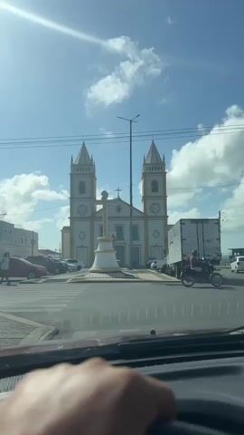 A wide shot shows a church with two towers and a cross in front of it, with a bright sunny sky and some clouds. Cars are parked on the side of the road, and a truck is driving down the street. The camera is inside a car, and a hand is visible on the steering wheel. The video is shot from the driver's perspective. The church appears to be white and yellow, and it has a clock on each tower. The cross in front of the church is white and gold. The cars are various colors, and the truck is white. The sky is blue, and there are a few white clouds. The sun is shining brightly. The video is taken from a slightly low angle, looking up at the church. The church appears to be in a town or city, as there are buildings on either side of it.