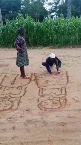 A young person wearing a white cap and pink shoes is crouching on the ground, drawing in the sand with their right hand. They are drawing on a large sand picture which appears to be two large figures. The figure on the right has a face drawn inside a circle with two eyes and a mouth. The figure on the left has a circle drawn inside it, with a few lines, and appears to be in an earlier stage of drawing. A woman stands to the left of the drawing, facing the young person, with her hands on her hips. She is wearing a dark patterned dress and orange sandals. The background consists of a dirt road with green corn stalks growing to the left. The sky is overcast.