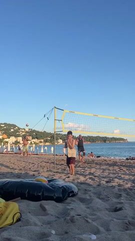 The video shows a beach scene with people playing volleyball. The sky is clear and blue, and the ocean is visible in the background. The sand is light brown, and there are a few beach chairs and umbrellas. In the foreground, there is a black inflatable object on the sand. Three people are playing volleyball. One person is serving the ball, and two others are on the opposite side of the net. The net is yellow and is set up on the beach. The people are wearing casual clothing. The scene appears to be during the daytime, possibly late afternoon, given the lighting.