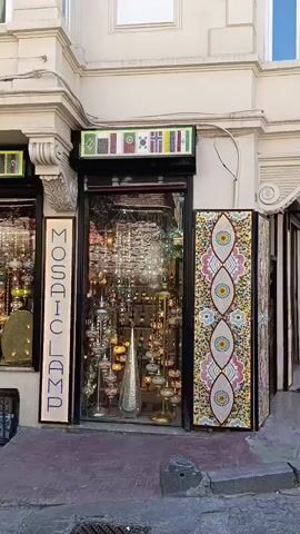 A camera pans across the storefront of a shop called "MOSAIC LAMP" displaying numerous mosaic lamps. The shop's facade is decorated with flags of various countries above the windows. On the left, a display case showcases more lamps. To the right of the entrance, a tall, narrow mosaic panel with an eye-like pattern is visible. In front of the shop, a family is walking by. A man pushes a stroller with a child inside, while a woman walks beside them. The street in front of the shop is paved with cobblestones, and the building's architecture appears to be old and ornate.