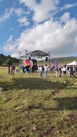 A group of people are gathered in a grassy outdoor area with a stage set up in the background. A car is being lifted by a crane. The sky is blue with scattered clouds.