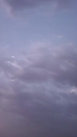 The video captures a time-lapse of a cloudy sky during what appears to be either dawn or dusk. The clouds are predominantly a muted purple and grey, with hints of lighter hues suggesting the presence of the sun either rising or setting. The camera angle is static, looking upwards at the sky, and no other elements are visible in the frame. The movement of the clouds indicates a gentle breeze. The overall atmosphere is serene and tranquil.