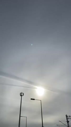 The video begins with a shot of a pale grey sky with a bright sun. In the lower portion of the frame, a cell tower and a lamppost are visible. The camera then pans and tilts upwards, showing a person wearing sunglasses and a grey beanie. The perspective shifts to an upside-down view, revealing a lamppost and a yellow car parked next to a fence. Beyond the fence, there are more lampposts and what appears to be a railway line with overhead wires and support structures. The ground is paved with interlocking bricks and bordered by grass. The video concludes with a closer view of the railway infrastructure.
