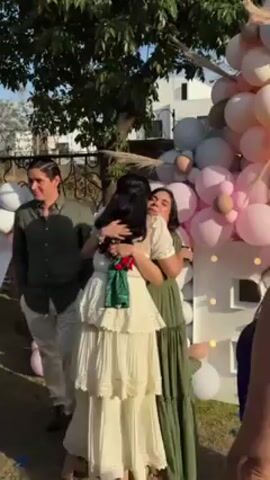 A baby shower is in progress outdoors. Three women and a man are visible, all interacting with each other in the frame. One woman wearing a tiered cream-colored dress is being hugged first by another woman in a green dress and then the man in an olive-colored shirt and white pants. A third woman in a sleeveless blue top is standing nearby, watching the interaction. Behind them is a decorative balloon arch in pink and white colors with a white cutout house. There is also a fence, a tree, and a white house visible in the background. The ground is grassy.