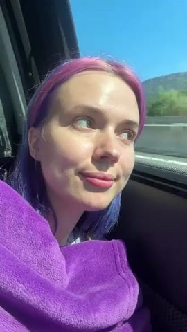 A person with purple and pink hair is sitting in a car, wrapped in a purple blanket, looking out the window. The sky is blue and there are mountains in the distance.