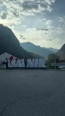 The video shows an “I LOVE RAJAVILI” sign in front of mountains. People are walking near the sign. A black SUV is parked nearby. The camera then pans to the right, showing the mountainous landscape with trees. The road and a metal guardrail are visible.