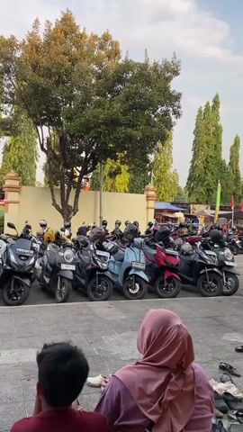 A group of people, including two in the foreground and others further back, are gathered in what appears to be a parking area. In the middle ground, a large number of motorcycles are parked in rows. A tall tree with green foliage is prominent behind the motorcycles. A beige-colored wall runs behind the parked vehicles, with trees visible on the other side. Two people, one wearing a hijab and a patterned outfit and another in a red shirt and jeans, are facing away from the camera, looking towards the motorcycles. A third person, wearing a pink hijab and purple top, is also seated facing away from the camera. The ground is paved, and there are several pairs of shoes scattered on the ground in the foreground. The sky is visible, with some clouds. The lighting suggests it is daytime, possibly late afternoon.