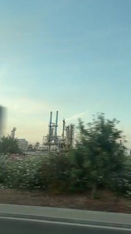 This video shows an outdoor view from a moving vehicle. The foreground features a gray road or sidewalk and a grassy embankment. In the mid-ground, there are trees and bushes, some appearing to be in motion due to the vehicle's movement. In the background, a large industrial facility with multiple tall structures, including chimneys, is visible against a pale blue sky with a few wispy clouds. A tall, thin pole with a light fixture at the top stands prominently in the middle of the frame in some shots. The overall impression is a landscape transitioning from natural elements to industrial infrastructure under a daytime sky.